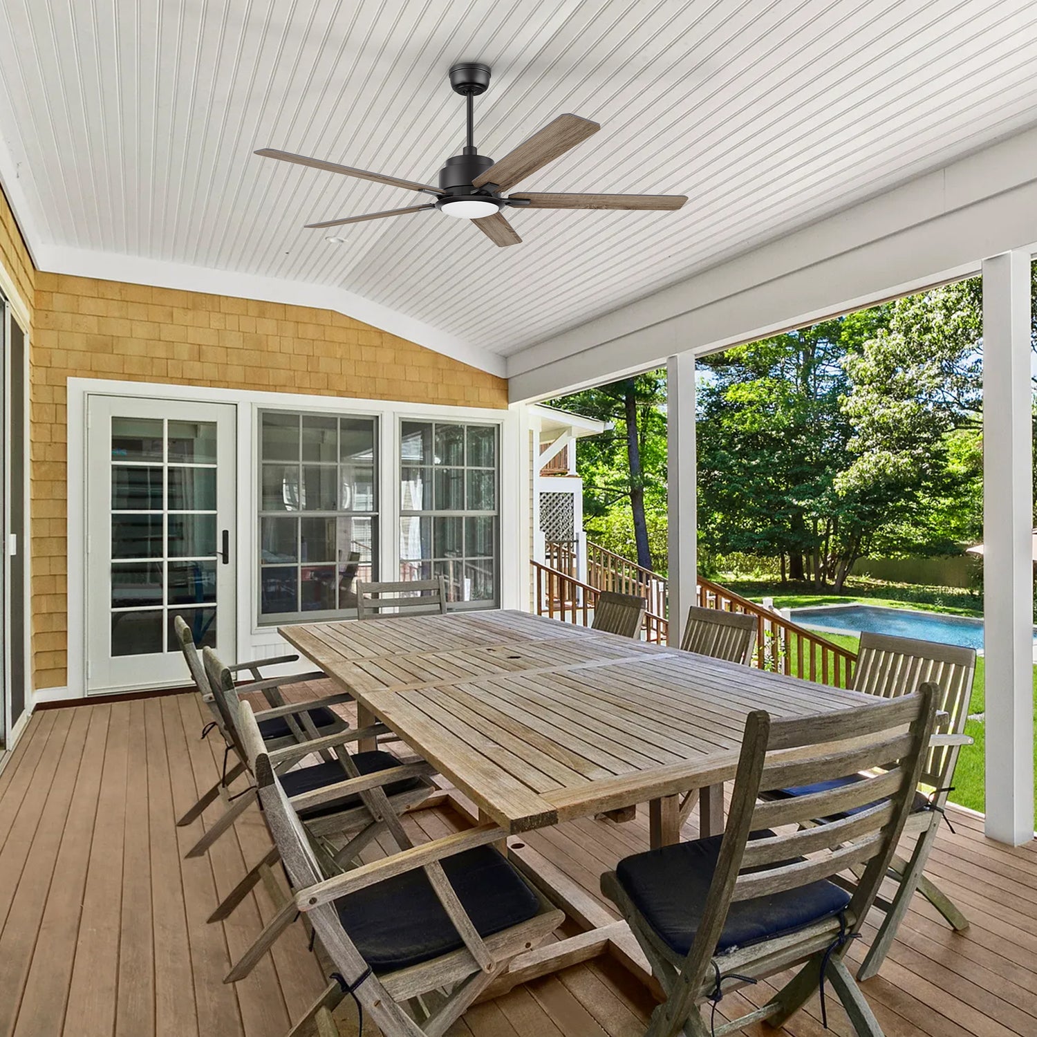 A spacious covered patio with a long wooden dining table and eight chairs, featuring the 56-inch Smafan Essex outdoor smart ceiling fan in walnut finish. The fan is mounted on a white paneled ceiling, providing cooling and lighting for the outdoor dining area. The space overlooks a backyard with lush greenery and a swimming pool. #color_walnut