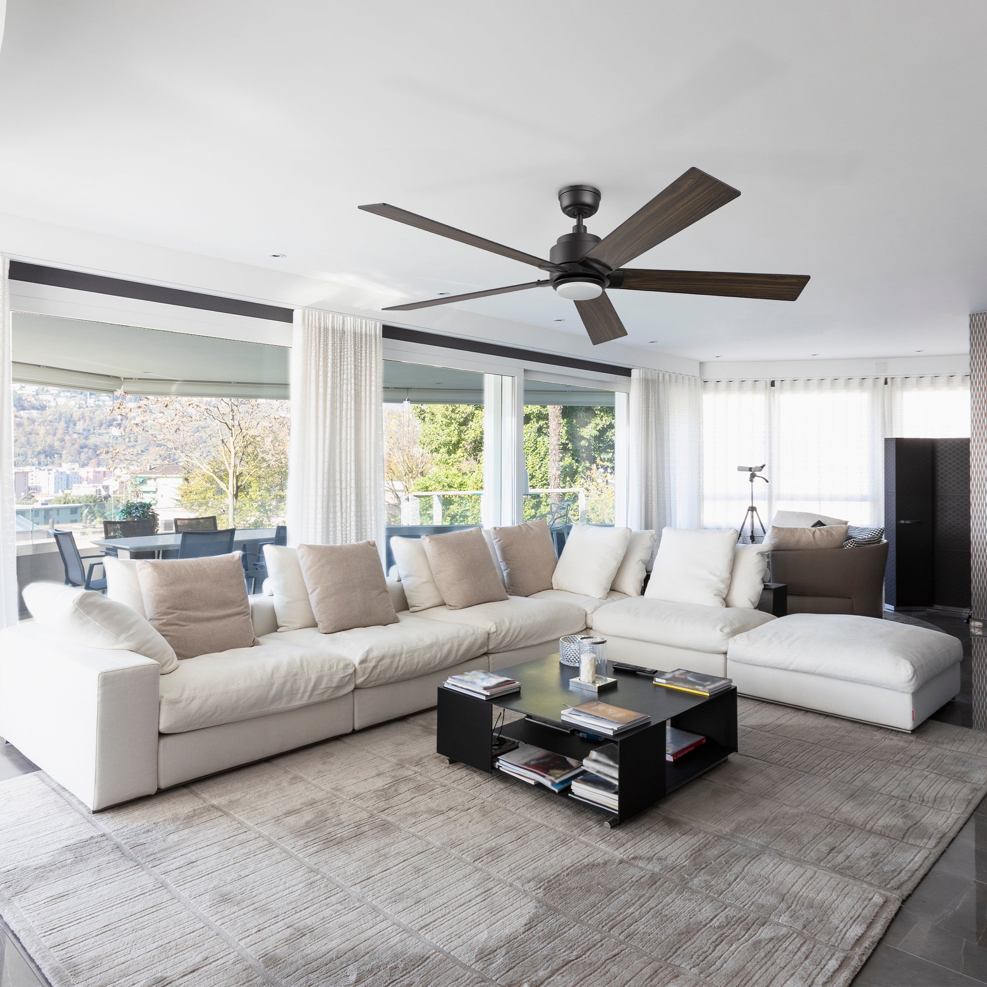 Spacious and bright modern living room with the Smafan Carro Aspen 60-inch smart outdoor ceiling fan, showcasing wood-tone blades and matte black finish, paired with a dimmable LED light. The room includes large windows, white curtains, and a comfortable white sectional sofa with a gray rug. 