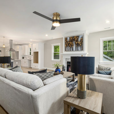 An open-concept living room and kitchen featuring a light grey sectional sofa with throw pillows, a wooden coffee table, and a matching side table with a gold-based lamp and dark blue shade. The room includes a fireplace with abstract artwork above it, and is illuminated by a flush mount ceiling fan in black and gold with an energy-efficient LED light kit, all styled in a modern farmhouse aesthetic with a neutral color palette. #color_black-gold