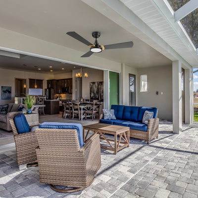 Modern covered patio with rattan furniture and blue cushions, featuring a black 52-inch Smafan Carro Viter smart outdoor ceiling fan with integrated LED light, installed overhead for comfortable airflow. The space opens into a stylish kitchen and dining area. #color_black-gold